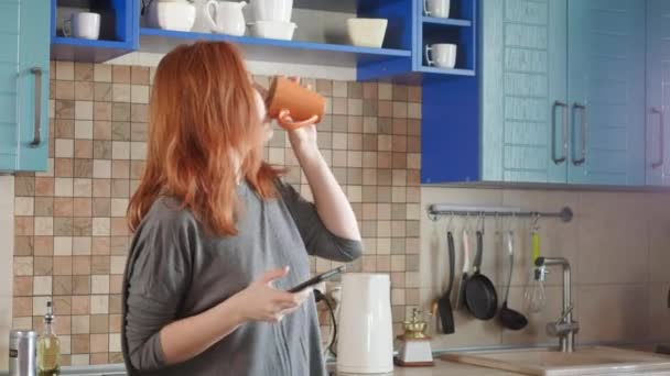 Danser amusant, écouter de la musique sur smartphone. Jolie fille aux cheveux roux boit le café du matin dans la cuisine de la maison. Jeune femme dansant et utilisant un téléphone portable. Femme rousse dansant dans la cuisine — Video