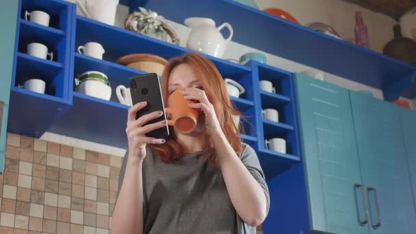 Zabawa taneczna, słuchanie muzyki na smartfonie. Atrakcyjna dziewczyna z rudymi włosami pije poranną kawę w domowej kuchni. Młoda kobieta tańczy i używa komórki. Rudowłosa kobieta tańczy w kuchni — Wideo stockowe