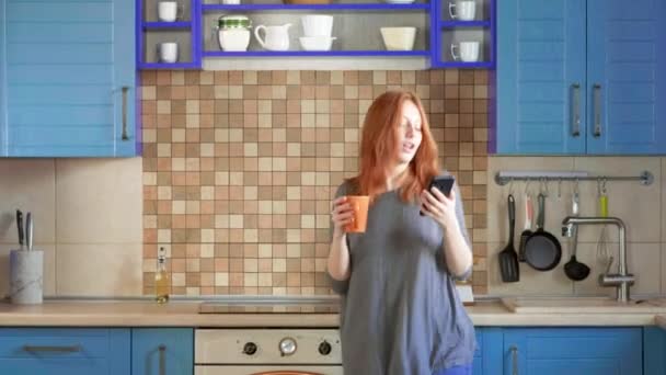 Bailando divertido, escuchando música en el smartphone. Chica atractiva con el pelo rojo bebe café de la mañana en la cocina del hogar. Joven mujer bailando y usando celular. Pelirroja bailando en la cocina — Vídeo de stock