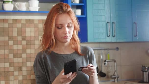 Menina atraente com cabelo vermelho faz uma compra na loja on-line usando um smartphone e um cartão de crédito. Compras passagens aéreas ou pedidos de entrega de alimentos. Serviço on-line de compras à distância de casa — Vídeo de Stock