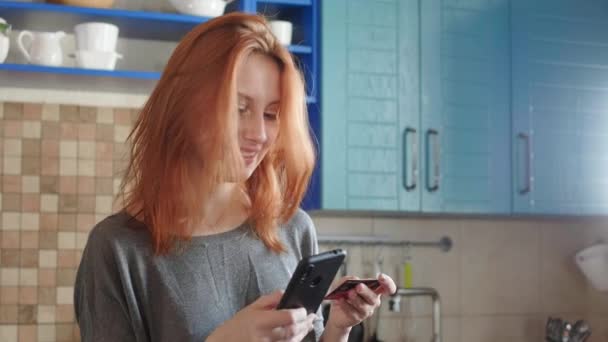 Attraktives Mädchen mit roten Haaren kauft mit Smartphone und Kreditkarte im Online-Shop ein. Kauft Flugtickets oder bestellt Essenslieferungen. Online-Service des Ferneinkaufs von zu Hause aus — Stockvideo