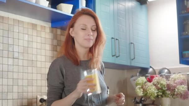 Menina bonita bebendo suco de laranja fresco de vidro. Mulher feliz bebendo bebida de frutas. Menina bonita em t-shirt de pé na cozinha. Nutrição saudável e natural. Café da manhã saudável no fim de semana — Vídeo de Stock