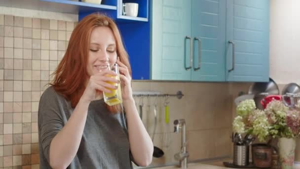 Atractiva joven pelirroja bebe jugo de naranja el fin de semana en la cocina. Utiliza un teléfono inteligente para hablar con los padres o amigos. Hablando por celular. Desayuno saludable — Vídeos de Stock