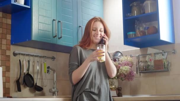 Attractive young redhead girl drinks orange juice at the weekend in the kitchen. Uses a smartphone to talk with parents or friends. Talking on a cell phone. Healthy breakfast — Stock Video