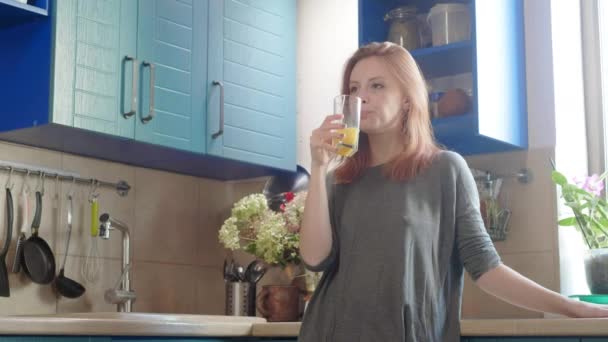 Красивая девушка пьет свежий апельсиновый сок из стекла. Счастливая женщина пьет фруктовый напиток. Красивая девушка в футболке, стоящая на кухне. Здоровое и естественное питание. Здоровый завтрак в выходные — стоковое видео