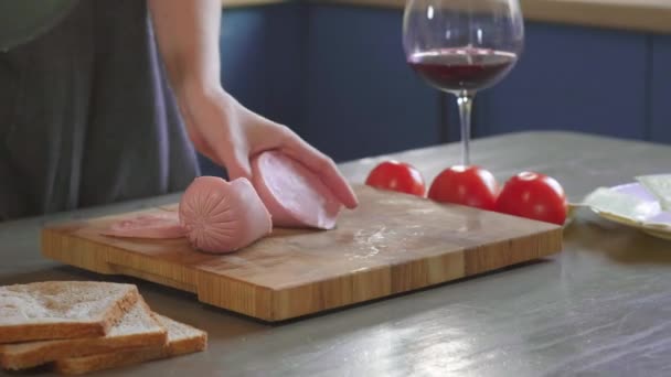 La mujer corta el sándwich del club por la mitad por el cuchillo agudo en la tabla de madera en la cocina, haciendo de la comida rápida en casa. Primer plano. Las niñas las manos cortan jamón o salchicha para un sándwich, desayuno — Vídeo de stock