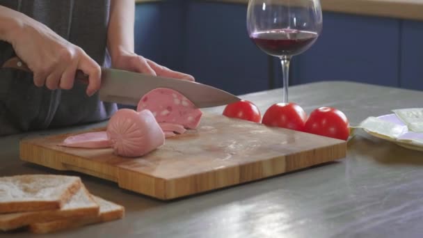 La mujer corta el sándwich del club por la mitad por el cuchillo agudo en la tabla de madera en la cocina, haciendo de la comida rápida en casa. Primer plano. Las niñas las manos cortan jamón o salchicha para un sándwich, desayuno — Vídeo de stock