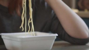 Çekici kızıl saçlı kız mutfakta makarna, makarna ve peynir yiyor. Abur cubur yiyor. Kötü bir yaşam tarzı. Telefonla konuşuyorum. Yemek hareket halinde. Özel plastik tabakta demlenmiş sarı ramen.