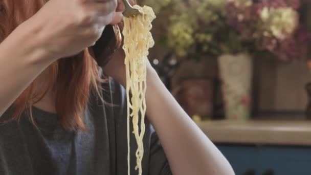 Приваблива руда дівчина їсть миттєву локшину пліт Мак і сир на кухні. Їсть нездорову їжу. Поганий спосіб життя. Говори по телефону. Їжа на ходу. Заварений жовтий рамен у спеціальній пластиковій тарілці — стокове відео