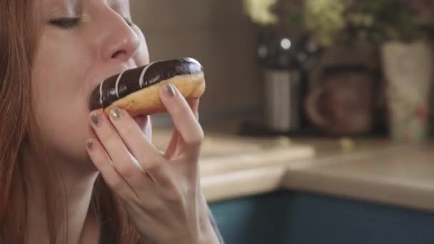 Menina com fome atraente ruiva come um donut no início da manhã no fim de semana na cozinha. Dieta não saudável, alimentos gordurosos, estilo de vida pobre em uma idade jovem — Vídeo de Stock