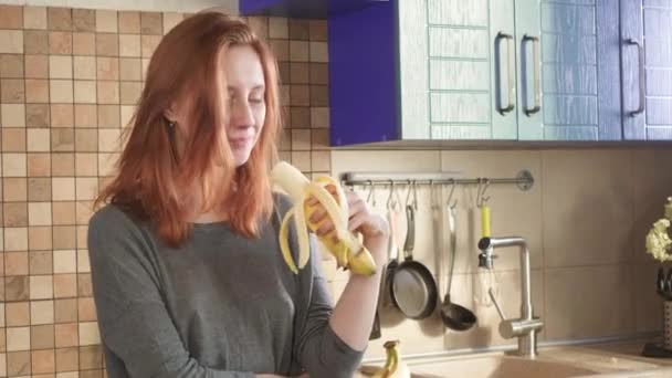 Atrakcyjna, głodna ruda dziewczyna zjada dużego banana wczesnym rankiem w weekend w kuchni. Czyści skórkę. Zdrowe odżywianie, właściwy styl życia. Śniadanie sportowe. — Wideo stockowe