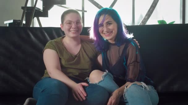 Two young lesbian girls sitting on the couch, looking at the camera, holding hands, hugging, cuddles, love, care, family idyll, smiling, happy. A girl with colored hair and a bald woman cuddling — Stock Video