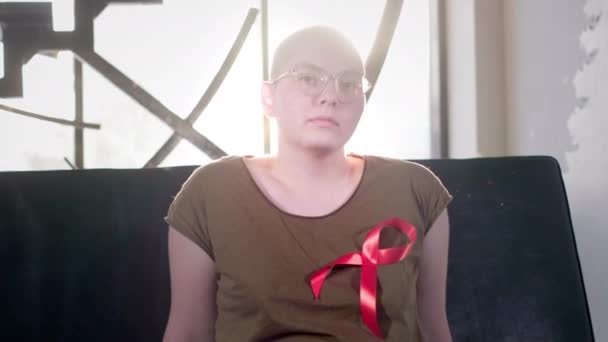 Łysa dziewczyna z AIDS lub Hiv siedzi w domu. Nosił czerwoną wstążkę. Rehabilitacja po chemioterapii. Wstążka uświadamiająca nowotwory. — Wideo stockowe
