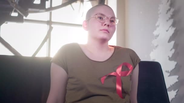 Łysa dziewczyna z AIDS lub Hiv siedzi w domu. Nosił czerwoną wstążkę. Rehabilitacja po chemioterapii. Wstążka uświadamiająca nowotwory. — Wideo stockowe