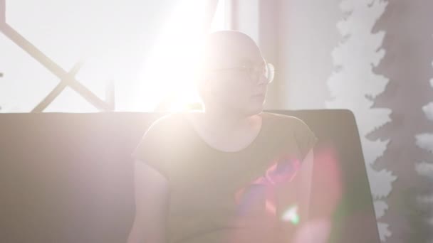 Una chica calva con SIDA o VIH está sentada en casa. Llevando una cinta roja. Rehabilitación después de la quimioterapia. Cinta de conocimiento del cáncer . — Vídeo de stock