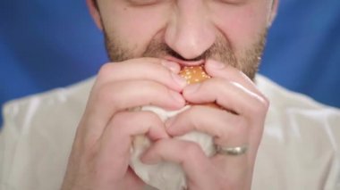 Yakın plan, sakallı bir adam hamburger yiyor. Tek eliyle çizburger tutuyormuş. Sağlıklı bir hayat sürmüyor, sağlık ve metabolizmayı bozuyor. Yanlış yemek.