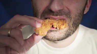 Yakın plan, sakallı bir adam tavuk bacağı yiyor. Tek eliyle nugget tutuyor. Sağlıklı bir hayat sürmüyor, sağlık ve metabolizmayı bozuyor. Yanlış yemek. Kanat ve patates kızartması.