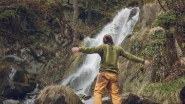 Genç bir erkek gezgin güzel bir şelalenin tadını çıkarıyor. Dağlarda yürüyüş. Yürüyüşçü şelaleye koşar, ellerini kaldırır. Yaşam tarzını seviyor. Kavram: Din, biyoloji, ekoloji.