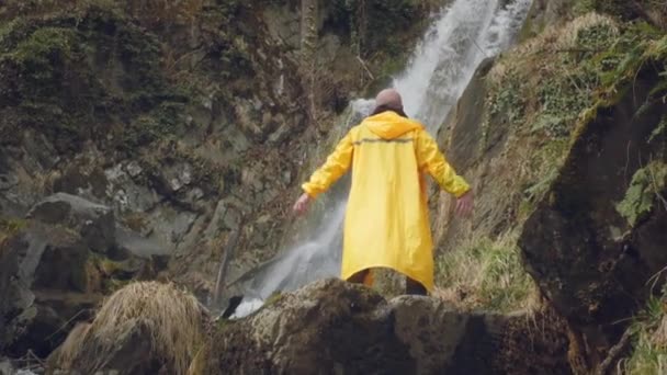 Young male traveler in a yellow raincoat enjoys a beautiful waterfall. Hiking in the mountains. The hiker runs to the waterfall, raises his hands up. Enjoys lifestyle. Concept: religion, bio, ecology. — Stock Video