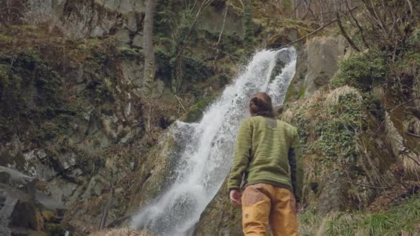 Joven viajero masculino en goza de una hermosa cascada. Senderismo en las montañas. El excursionista corre hacia la cascada, levanta las manos. Disfruta del estilo de vida. Concepto: religión, bio, ecología. Movimiento lento . — Vídeo de stock