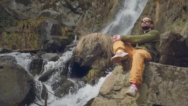 Jeune voyageur masculin assis jouit d'une belle cascade. Randonnée dans les montagnes. Un randonneur s'assied à une cascade, lève les mains. Apprécie le mode de vie dans les Alpes françaises . — Video