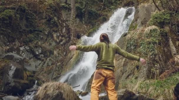 Joven viajero masculino en goza de una hermosa cascada. Senderismo en las montañas. El excursionista corre hacia la cascada, levanta las manos. Disfruta del estilo de vida. Concepto: religión, bio, ecología . — Vídeos de Stock