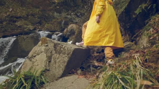 Young male traveler in a yellow raincoat enjoys a beautiful waterfall. Hiking in the mountains. The hiker runs to the waterfall, raises his hands up. Lifestyle in slow motion. Concept: religion, bio. — Stock Video