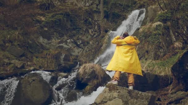 Young male traveler in a yellow raincoat enjoys a beautiful waterfall. Hiking in the mountains. The hiker runs to the waterfall, raises his hands up. Lifestyle in slow motion. Concept: religion, bio. — Stock Video