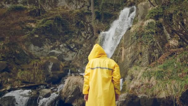 Young male traveler in a yellow raincoat enjoys a beautiful waterfall. Hiking in the mountains. The hiker runs to the waterfall, raises his hands up. Lifestyle in slow motion. Concept: religion, bio. — Stock Video