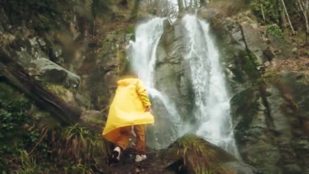 Jovem viajante do sexo masculino em uma capa de chuva amarela goza de uma bela cachoeira. Caminhadas nas montanhas. O caminhante corre para a cachoeira, levanta as mãos. Estilo de vida em câmara lenta. Conceito: religião, bio . — Vídeo de Stock