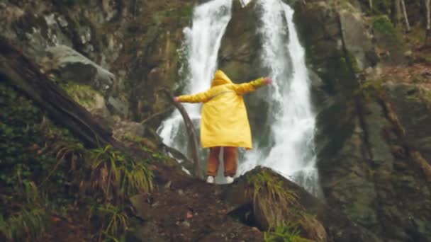 Young male traveler in a yellow raincoat enjoys a beautiful waterfall. Hiking in the mountains. The hiker runs to the waterfall, raises his hands up. Lifestyle in slow motion. Concept: religion, bio. — Stock Video