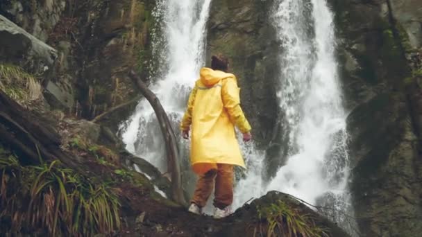 穿着黄色雨衣的年轻男性旅行者喜欢美丽的瀑布。在山上远足。徒步旅行者跑到瀑布边，举起双手。慢动作的生活方式。概念：宗教、生物. — 图库视频影像