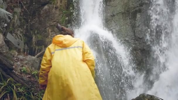 Joven viajero masculino en un impermeable amarillo disfruta de una hermosa cascada. Senderismo en las montañas. El excursionista corre hacia la cascada, levanta las manos. Estilo de vida en cámara lenta. Concepto: religión, bio . — Vídeo de stock