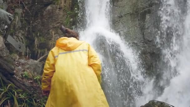 Giovane viaggiatore maschio in un impermeabile giallo gode di una bella cascata. Escursioni in montagna. L'escursionista corre verso la cascata, alza le mani. Stile di vita al rallentatore. Concetto: religione, bio . — Video Stock