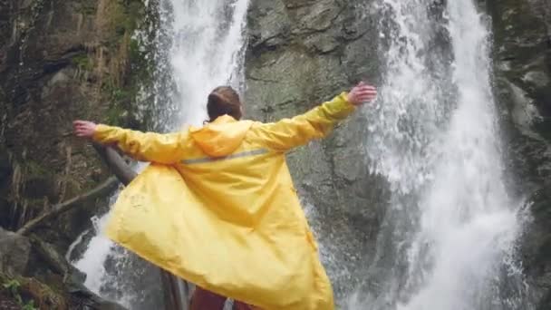 Joven viajero masculino en un impermeable amarillo disfruta de una hermosa cascada. Senderismo en las montañas. El excursionista corre hacia la cascada, levanta las manos. Estilo de vida en cámara lenta. Concepto: religión, bio . — Vídeos de Stock