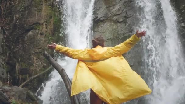 穿着黄色雨衣的年轻男性旅行者喜欢美丽的瀑布。在山上远足。徒步旅行者跑到瀑布边，举起双手。慢动作的生活方式。概念：宗教、生物. — 图库视频影像