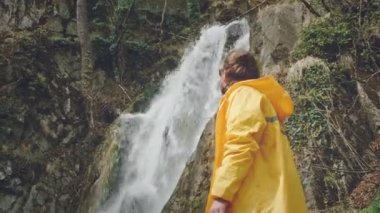 Sarı yağmurluk giyen genç bir erkek gezgin güzel bir şelalenin tadını çıkarıyor. Dağlarda yürüyüş. Yürüyüşçü şelaleye koşar, ellerini kaldırır. Yaşam tarzını seviyor. Kavram: Din, biyoloji, ekoloji.