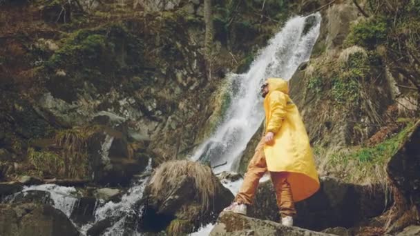 穿着黄色雨衣的年轻男性旅行者喜欢美丽的瀑布。在山上远足。徒步旅行者跑到瀑布边，举起双手。享受生活的乐趣。概念：宗教、生物、生态. — 图库视频影像