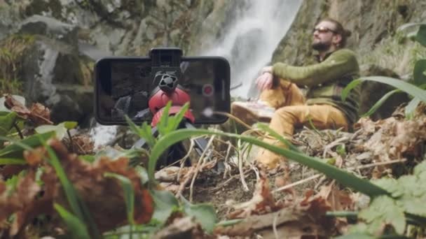 Junge männliche Reisende Blogger im Sitzen genießt einen schönen Wasserfall. Wandern in den Bergen. Ein Wanderer sitzt an einem Wasserfall, nimmt ein Video mit dem Handy auf. Genießt Lifestyle in den französischen Alpen. — Stockvideo