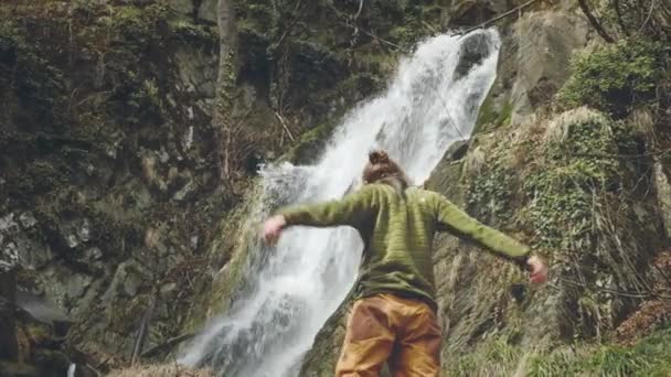 美しい滝を楽しむ若い男性旅行者。山の中でハイキング。ハイカーは滝に走り、手を挙げる。ライフスタイルを楽しむ。概念:宗教、生物、生態学. — ストック動画
