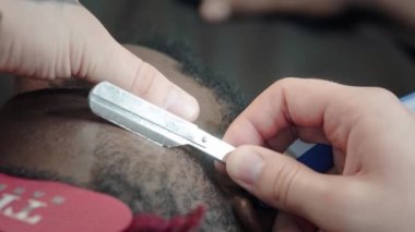 Yakın plan: bir kuaför bir Afrikalı Amerikalının saçlarını tıraş eder. Şık bir saç stili yapıyor, tıraş olmak için tehlikeli bir jilet kullanıyor. Saç stilini yumuşatır.