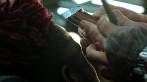 Gros plan : un coiffeur se rase les cheveux sur la tête d'un afro-américain. Fait une coiffure élégante, utilise un trimer de rasage électrique. Les cadenas sont rouges sur la tête . — Video