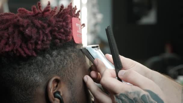 클로즈업: 한 미용사가 아프리카 계 미국인의 머리를 깎아 냅니다. 멋진 헤어 스타일을 만들고 전기 샤워기를 사용 합니다. 잠금장치가 머리에 붉게 쓰여 있다. — 비디오