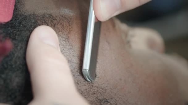 Close-up: een kapper scheert het haar af op het hoofd van een Afro-Amerikaan. Maakt een stijlvolle kapsel, maakt gebruik van een gevaarlijk scheermes voor het scheren. Verzacht de contour van het kapsel — Stockvideo