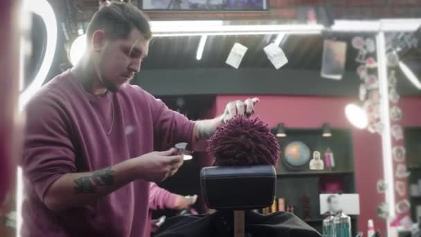 Primer plano: un peluquero se afeita el pelo en la cabeza de un afroamericano. Hace un peinado elegante, utiliza un trimer de afeitado eléctrico. Las rastas son rojas en la cabeza. . — Vídeo de stock