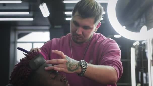 Zbliżenie: fryzjer goli włosy na głowie Afroamerykanina. Robi stylową fryzurę, używa niebezpiecznej brzytwy do golenia. Wygładza kontur fryzury — Wideo stockowe