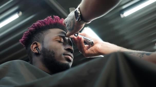 Nahaufnahme: Ein Friseur rasiert einem Afroamerikaner die Haare auf dem Kopf ab. Macht eine stylische Frisur, verwendet einen elektrischen Rasierapparat. Dreadlocks sind rot auf dem Kopf. — Stockvideo