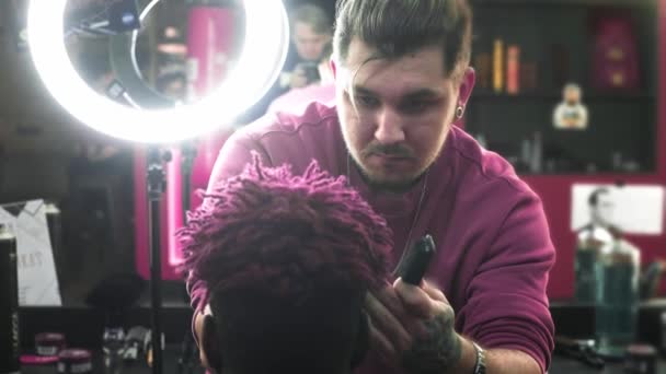 Nahaufnahme: Ein Friseur rasiert einem Afroamerikaner die Haare auf dem Kopf ab. Macht eine stylische Frisur, verwendet einen elektrischen Rasierapparat. Dreadlocks sind rot auf dem Kopf. — Stockvideo