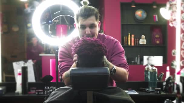 Nahaufnahme: Ein Friseur rasiert einem Afroamerikaner die Haare auf dem Kopf ab. Macht eine stylische Frisur, verwendet einen elektrischen Rasierapparat. Dreadlocks sind rot auf dem Kopf. — Stockvideo