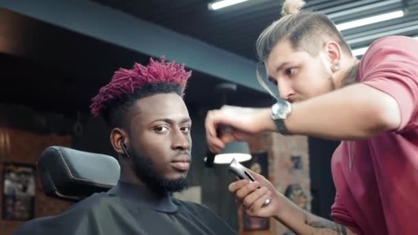 Primer plano: un peluquero se afeita el pelo en la cabeza de un afroamericano. Hace un peinado elegante, utiliza un trimer de afeitado eléctrico. Las rastas son rojas en la cabeza. . — Vídeos de Stock